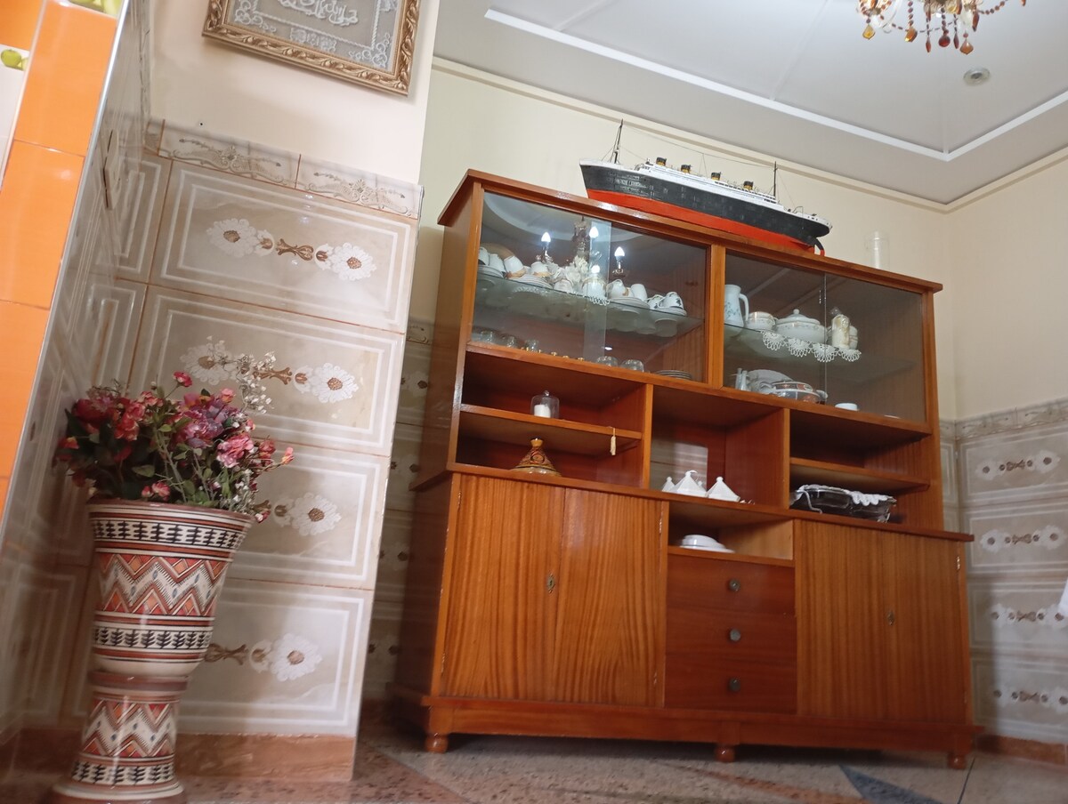 A wooden display cabinet is positioned against a patterned wall, showcasing delicate china and decorative objects. A model ship rests atop the cabinet, while a decorative vase filled with artificial flowers adds a touch of color to the corner.