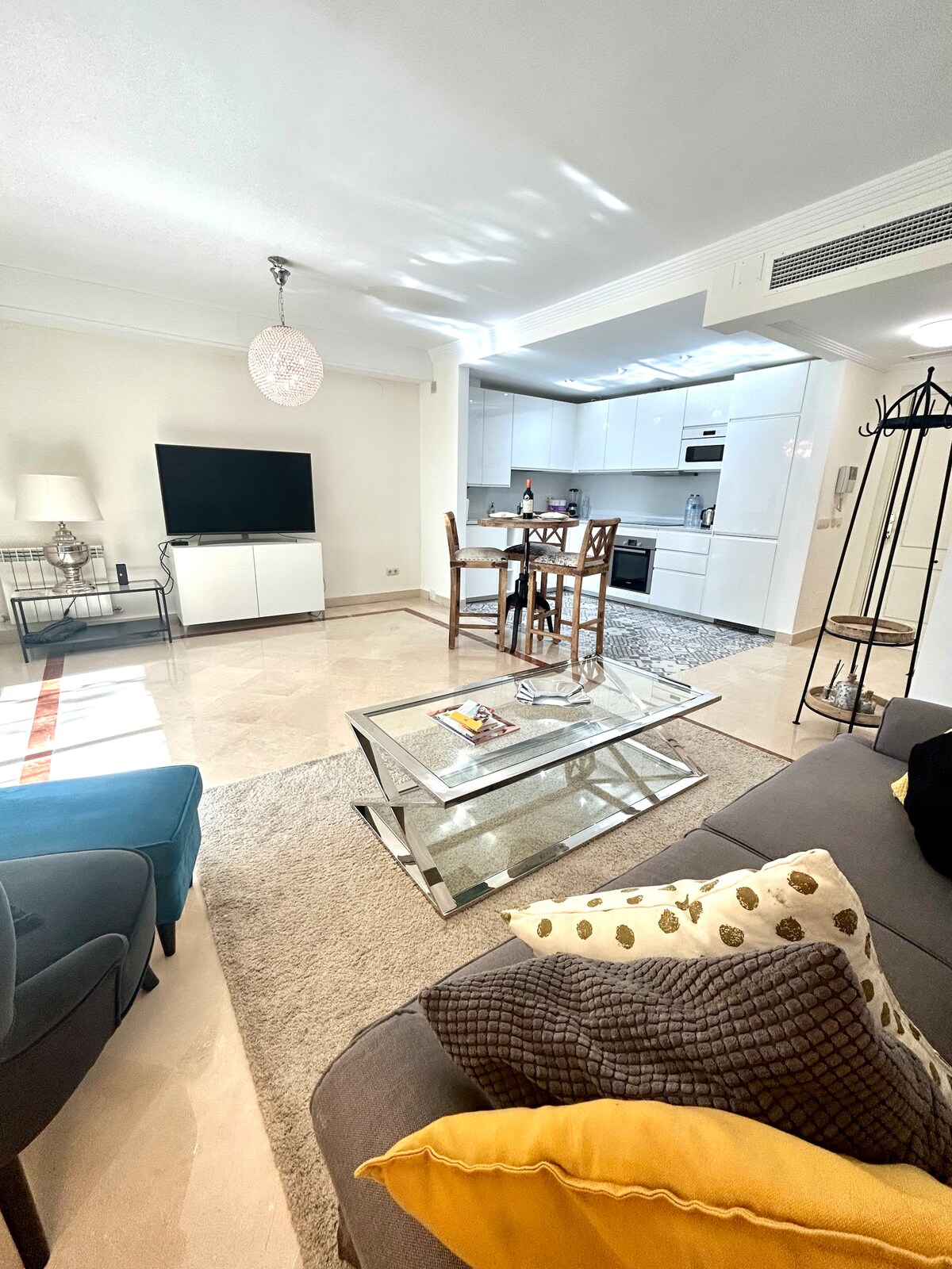 The living area features a light and airy design with a mix of elegant furnishings. A comfortable sofa is adorned with yellow and patterned pillows, facing a flat-screen television. A dining table with high chairs is positioned nearby, while a soft rug adds warmth to the space.