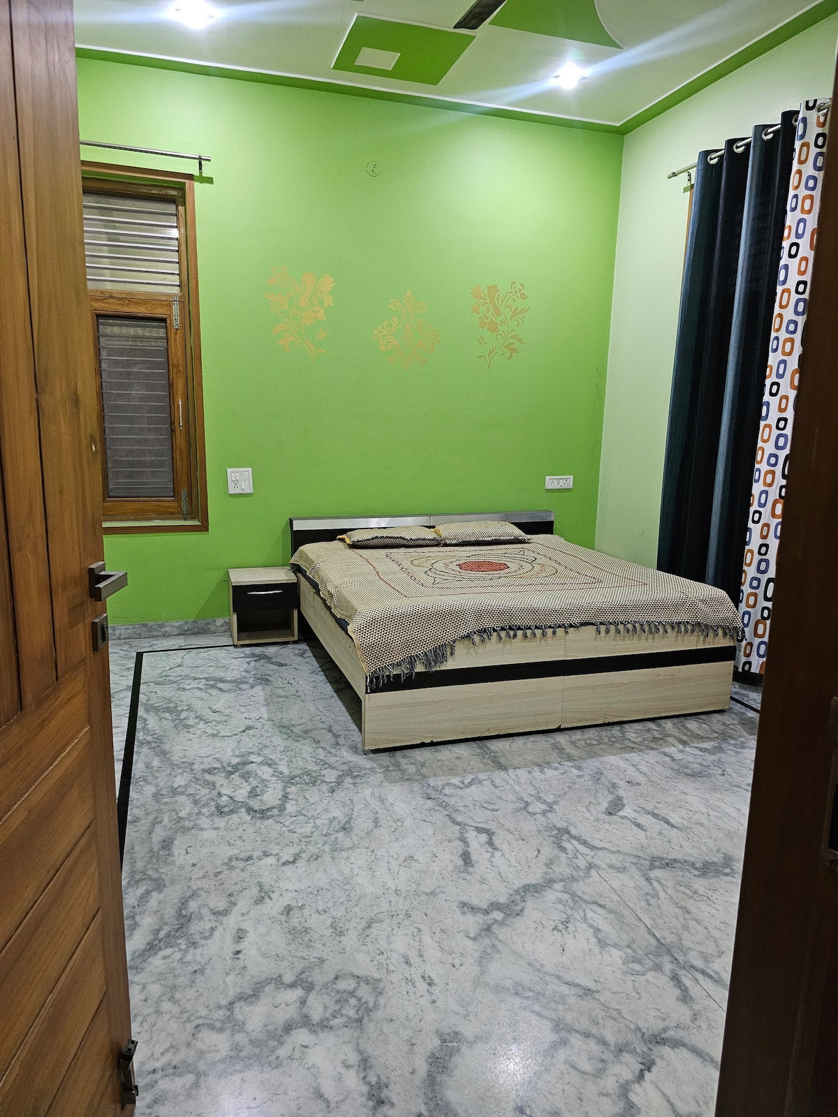 The bedroom features green walls complemented by patterned curtains. A spacious bed is adorned with a light-colored blanket and an accent piece. A nightstand sits beside the bed, and a large window allows natural light to brighten the room.