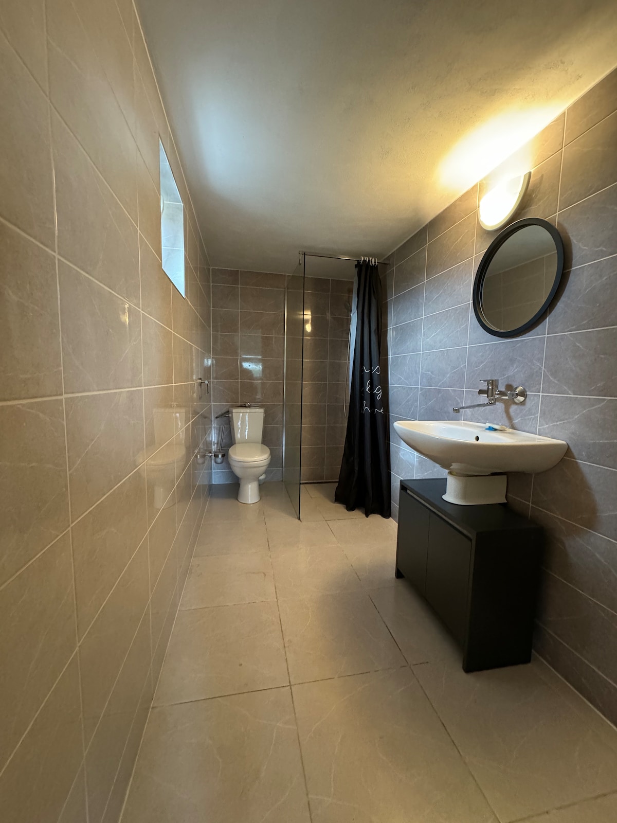 A clean and modern bathroom features tiled walls and floors in neutral tones. A shower is enclosed with a glass panel, and a circular mirror is mounted above a basin. A dark cabinet provides storage, while natural light enters through a small window.