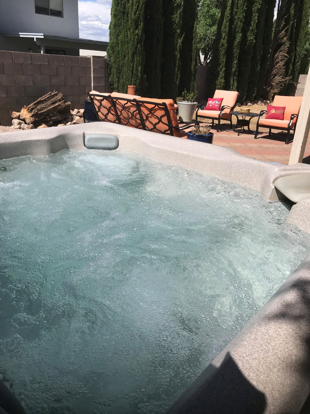 A bubbling hot tub is set in a private outdoor space, reflecting sunlight. Comfortable lounge chairs are arranged nearby on a brick patio, with decorative pillows. Tall privacy hedges contribute to the serene atmosphere of the area.