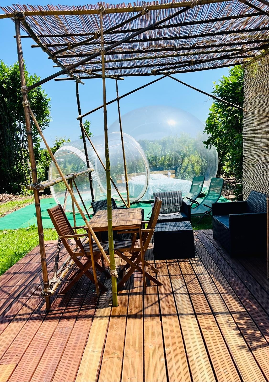 An outdoor dining area is set under a bamboo pergola, featuring a wooden table surrounded by folding chairs. Nearby, transparent bubbles are visible, with a seating arrangement that includes a dark outdoor sofa. Lush greenery and a well-maintained garden enhance the serene environment.