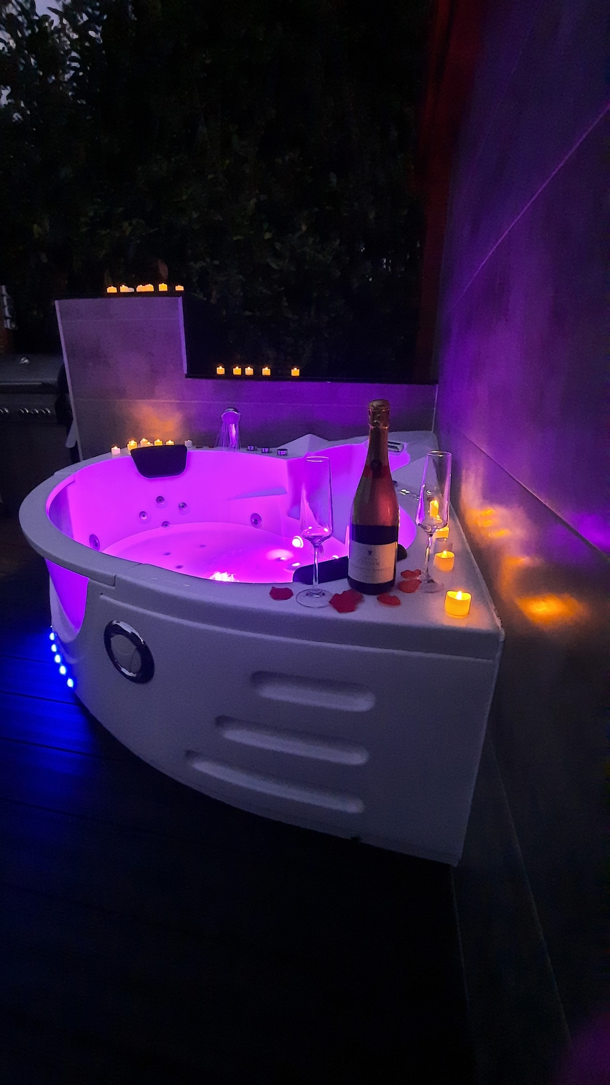 A private hot tub is illuminated with soft purple lighting, surrounded by flickering candles. Glasses and a bottle of champagne are positioned nearby, complementing the tranquil setting for relaxation.