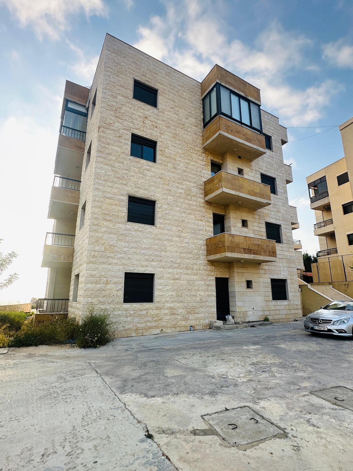 A modern multi-story building is presented, featuring a light-colored stone façade and multiple balconies with glass railings. Surrounding greenery is visible, complemented by a paved parking area in the foreground. The sky above is bright with scattered clouds.