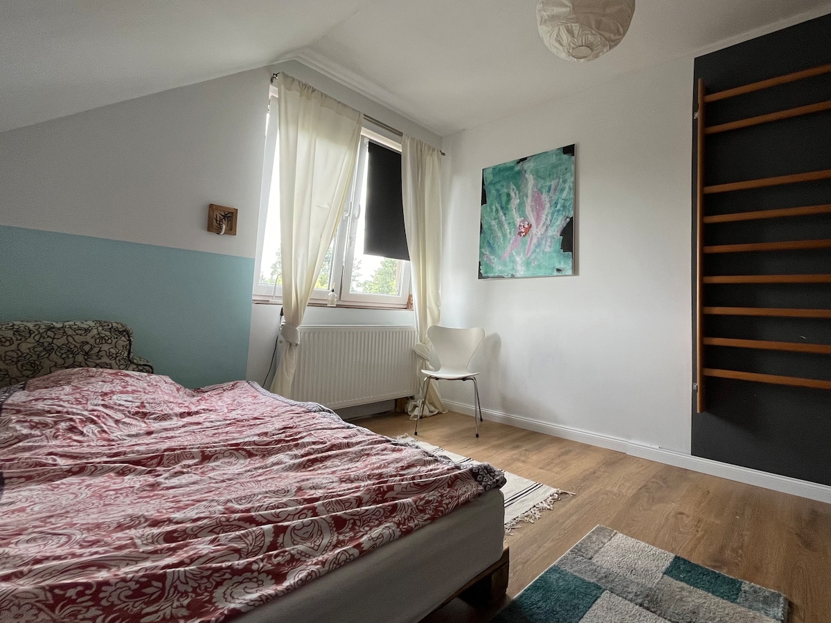 A simple bedroom is presented with a spacious bed covered by a patterned blanket. Natural light enters through a window, while a minimalist chair and a colorful artwork are visible. A narrow shelf is mounted on the wall, providing additional storage options.