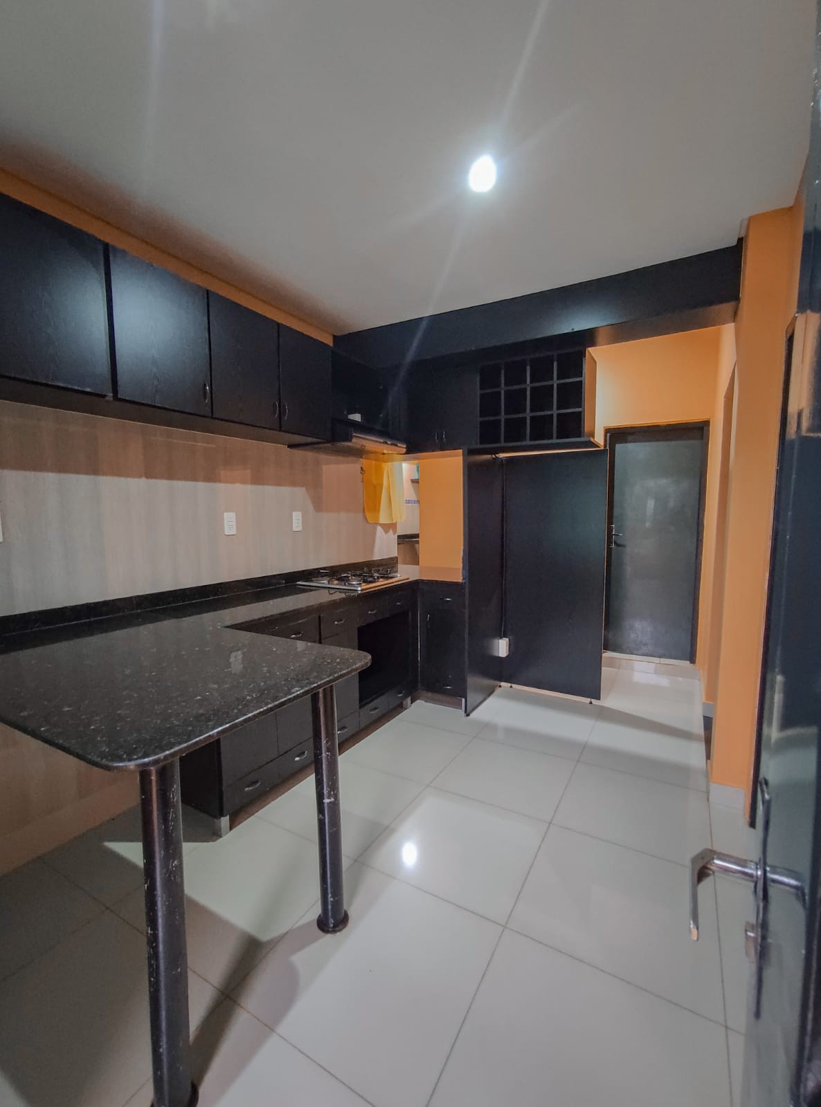 The kitchen features dark cabinetry and a black countertop with a modern stove. Light-colored walls contrast with the cabinetry, while the floor showcases glossy tiles. A small table provides additional workspace, and a wine rack is visible in the corner.