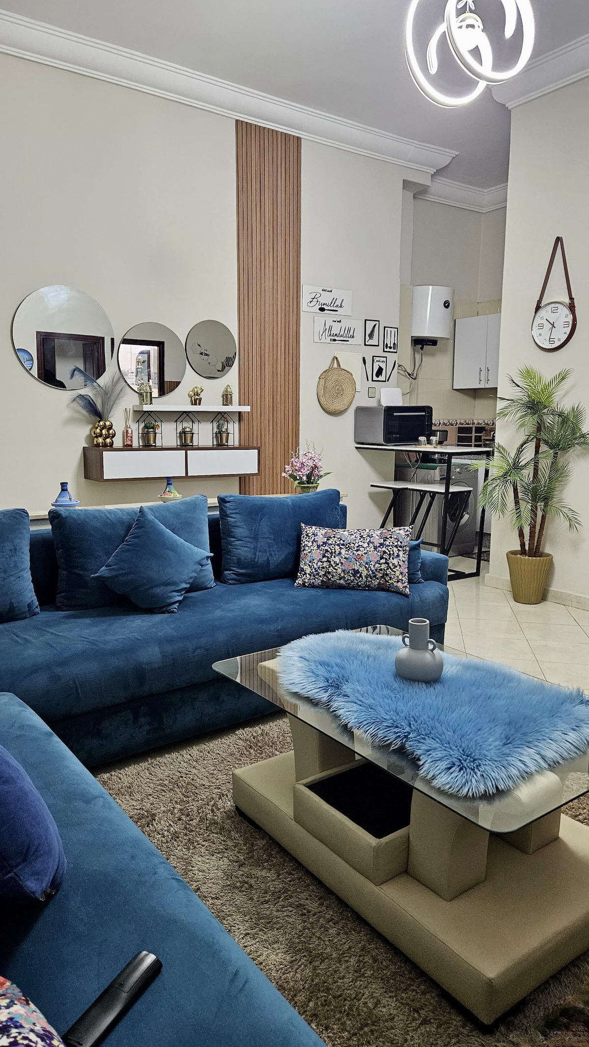 A spacious living room features soft blue sofas adorned with throw pillows arranged around a modern coffee table. A plush area rug lies beneath, while decorative mirrors enhance the ambiance. A small desk is visible in the background, alongside a subtle indoor plant for added decor.