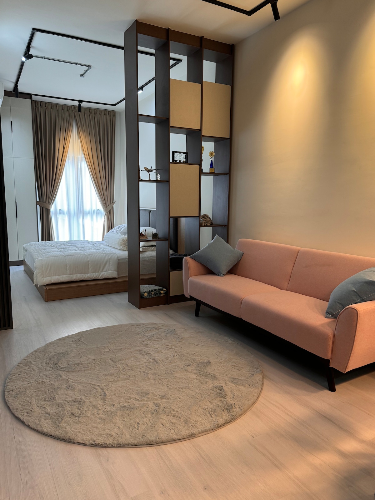 A cozy seating area features a soft pink sofa and a round area rug, complementing the light wood flooring. A partial room divider reveals a well-lit sleeping space with a spacious bed, framed by soft drapes and natural light filtering through a window.