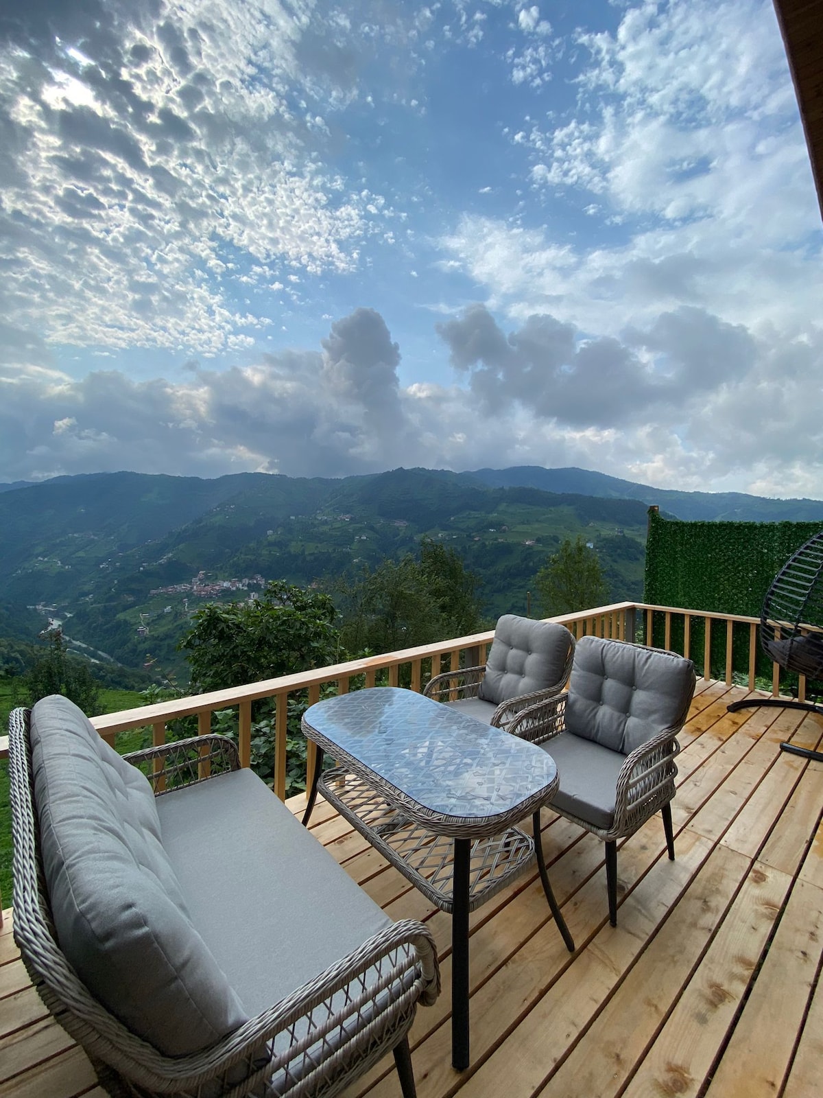 A spacious outdoor deck is furnished with a glass table and comfortable seating for four. Lush green mountains and a dramatic sky are visible in the background, contributing to a serene outdoor experience.