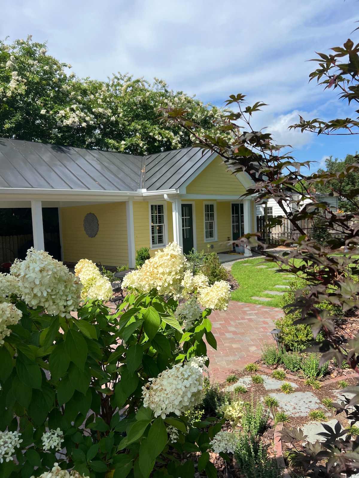 A charming gatehouse sits behind a yellow main house, framed by lush greenery and blooming hydrangeas. The exterior features a metal roof and multiple windows, allowing natural light to enhance the inviting facade. Paved pathways and a well-maintained yard invite relaxation in the garden.