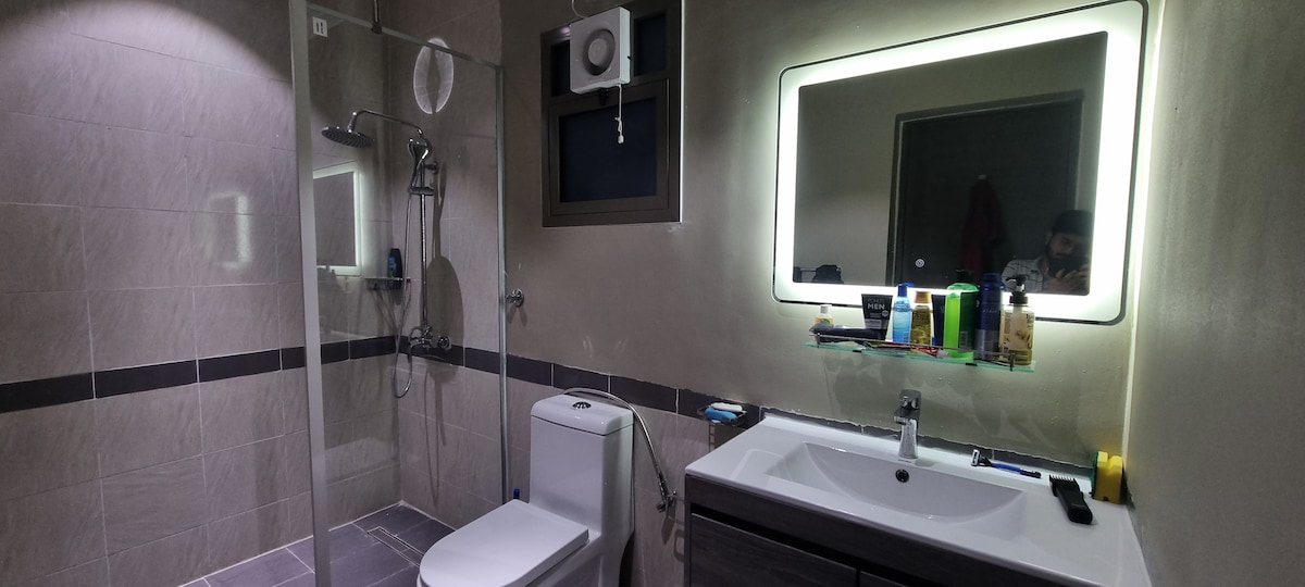 A modern bathroom is displayed, featuring a glass shower enclosure and a sleek vanity with a large illuminated mirror. Neutral-toned tiles line the walls, and essential toiletries are arranged on a shelf above the sink, providing a functional and clean environment.