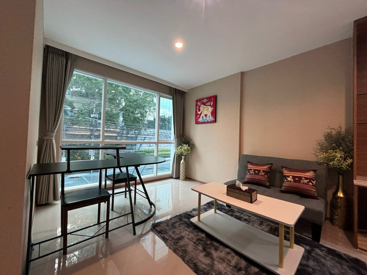 A contemporary living space features a large window allowing natural light to fill the room. A dining table is positioned beside the window, accompanied by stylish chairs. A modern coffee table sits in front of a cozy seating area adorned with decorative cushions, enhancing the room's aesthetic.