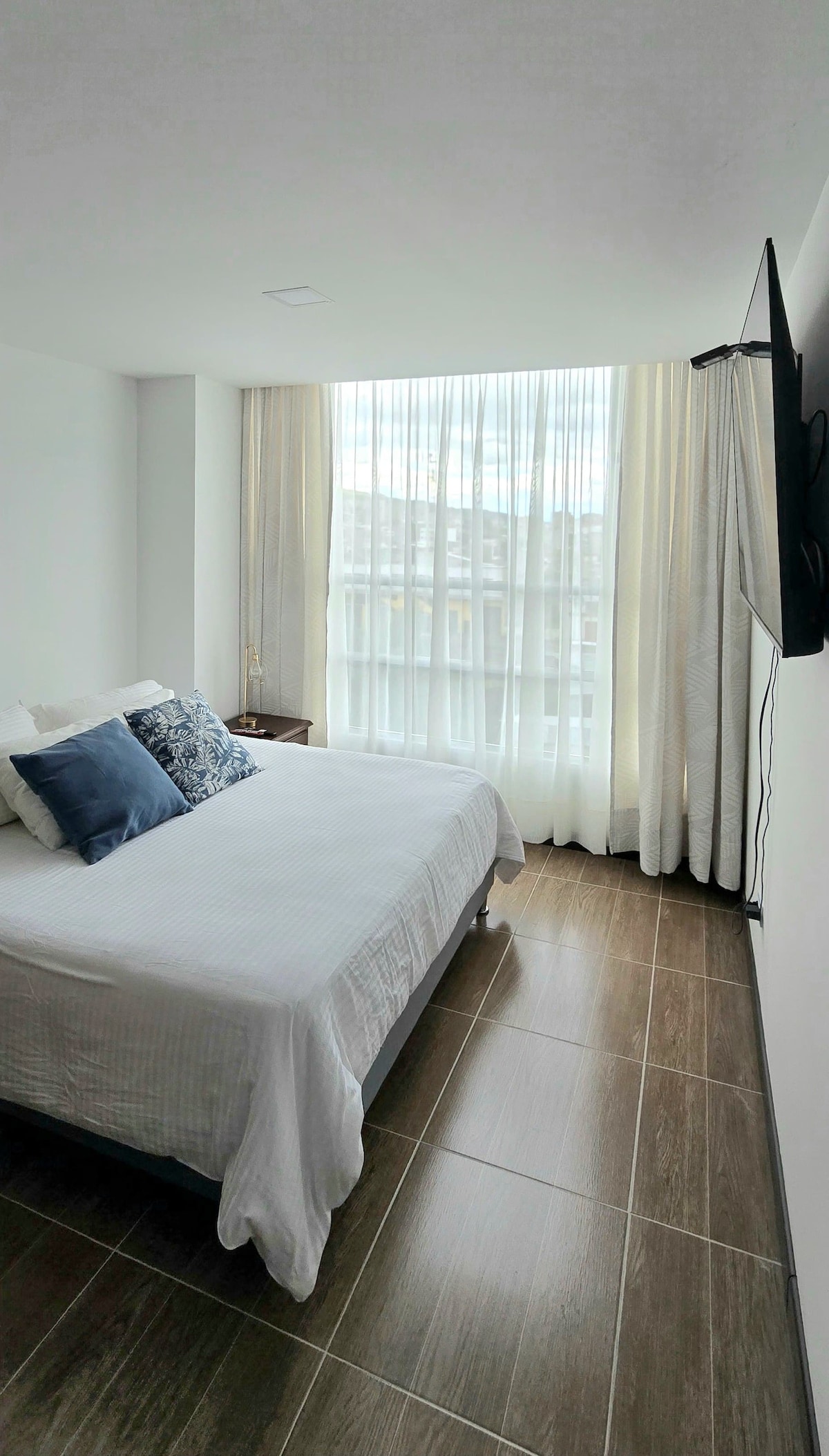 A well-lit bedroom is furnished with a spacious bed, adorned with decorative pillows. Sheer curtains allow natural light to fill the space, while a television is mounted on the wall. Neutral tones and tile flooring contribute to a modern atmosphere.