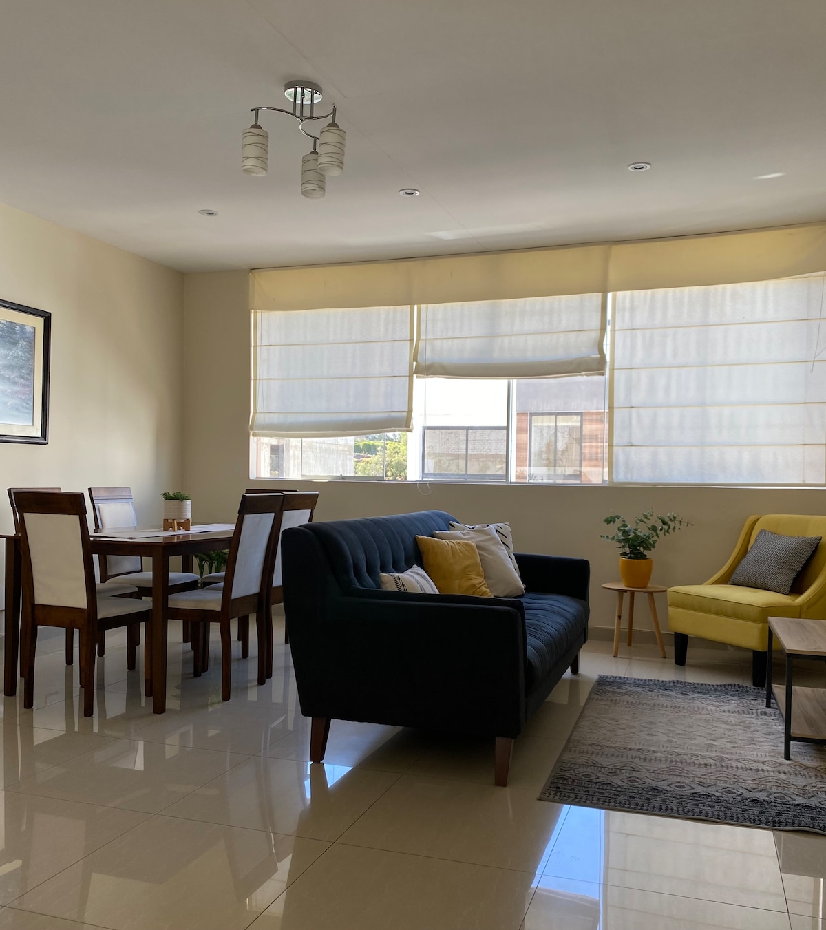 A bright living area showcases a dark sofa paired with a yellow armchair. A small dining table for four is situated near the window, which is adorned with light-filtering shades. The floor is tiled, and a subtle area rug adds comfort to the space.