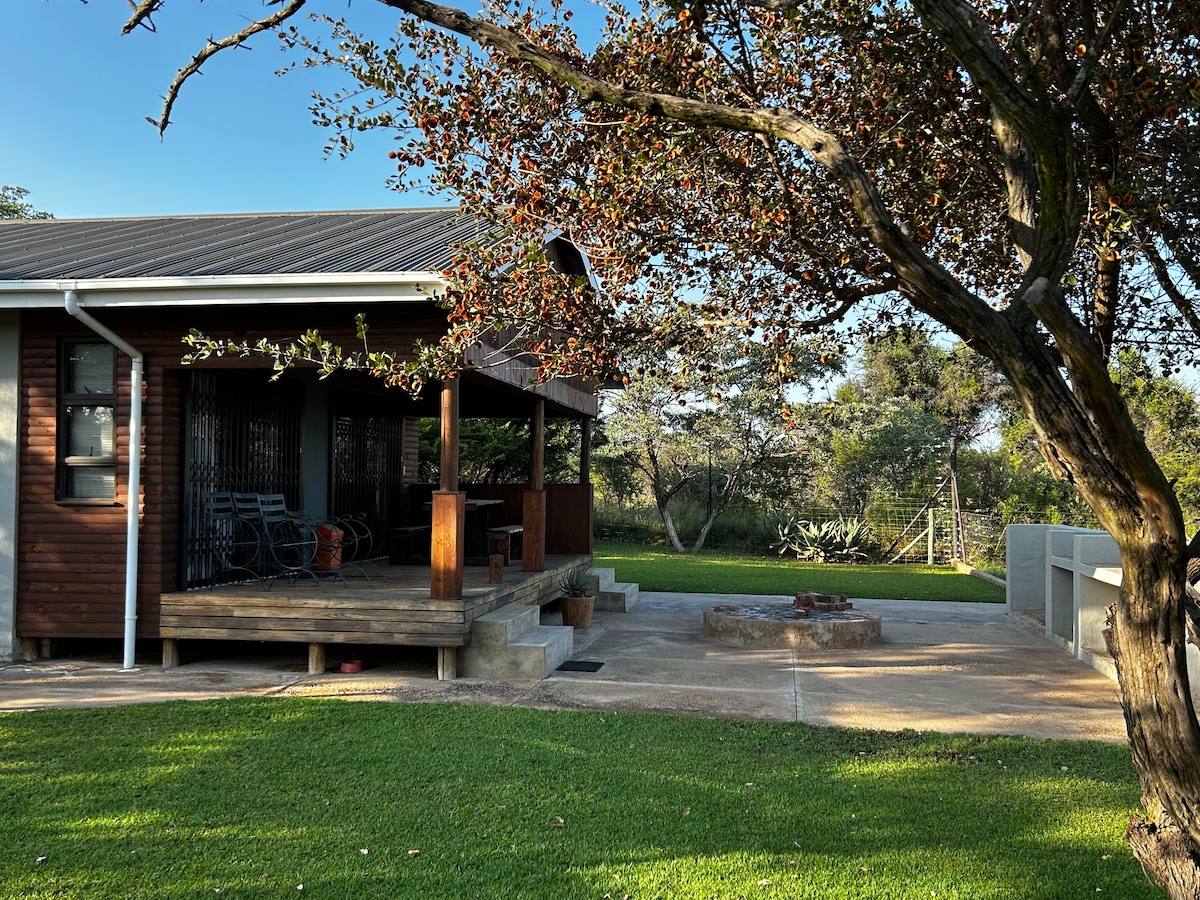 A wooden cabin features a spacious deck equipped with seating, offering a view of the surrounding bush. A fire pit is positioned on a stone patio, framed by a tree with flowering branches. The well-kept lawn extends toward the natural landscape.