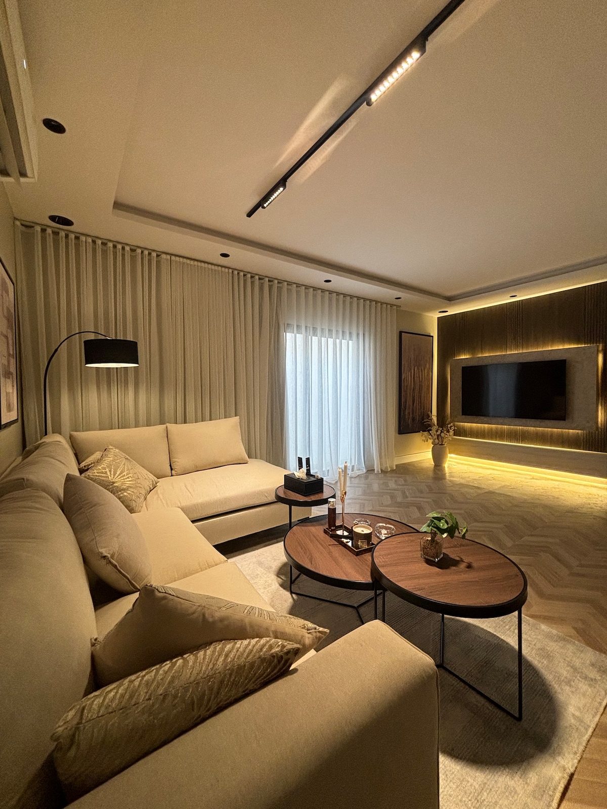 A modern living area features a spacious sectional sofa in neutral tones, accompanied by a pair of round coffee tables. Soft lighting accentuates textured curtains and a wall-mounted television, while a small plant adds a touch of greenery to the inviting space.