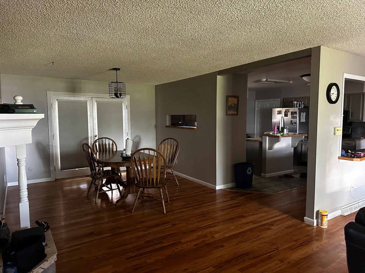 An open dining area featuring a wooden table surrounded by six matching chairs sits beside a partition leading to the kitchen. Natural light flows through large windows, highlighting the smooth wooden flooring. A fireplace adds a cozy touch to the space.