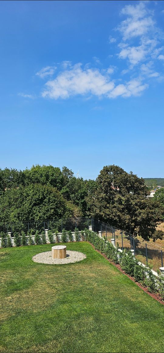 A serene outdoor space features a well-maintained lawn, surrounded by a fence and lush greenery. A circular stone arrangement sits at the center, framed by trees and a clear blue sky overhead, creating a tranquil environment for relaxation.