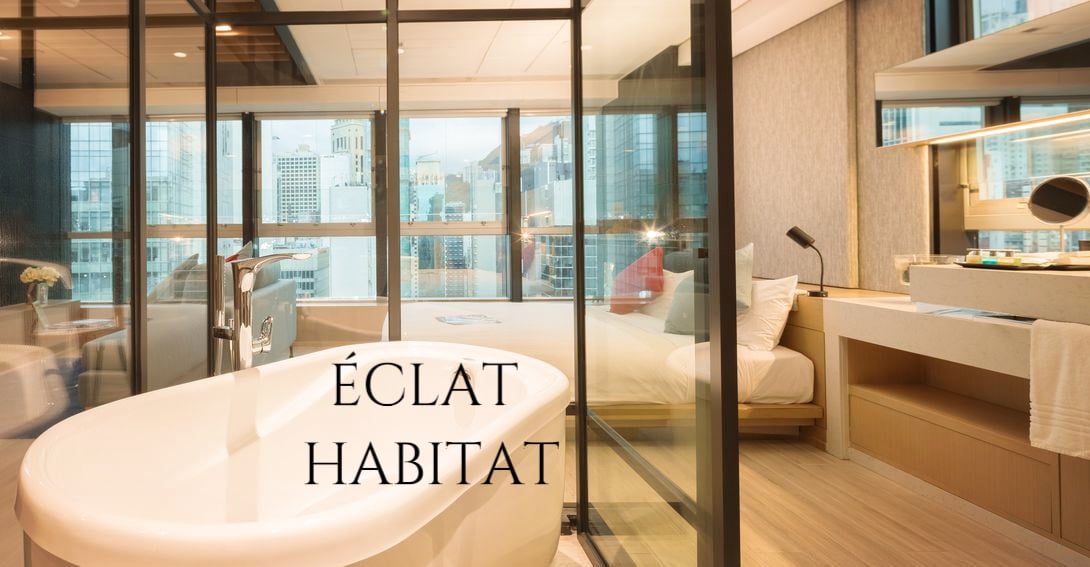 A modern bathroom featuring a freestanding bathtub is situated beside a large window, providing views of the city skyline. The glass-enclosed layout separates the bathing area from a well-appointed sleeping space, illuminated by natural light. Elegant furnishings add to the serene ambiance.