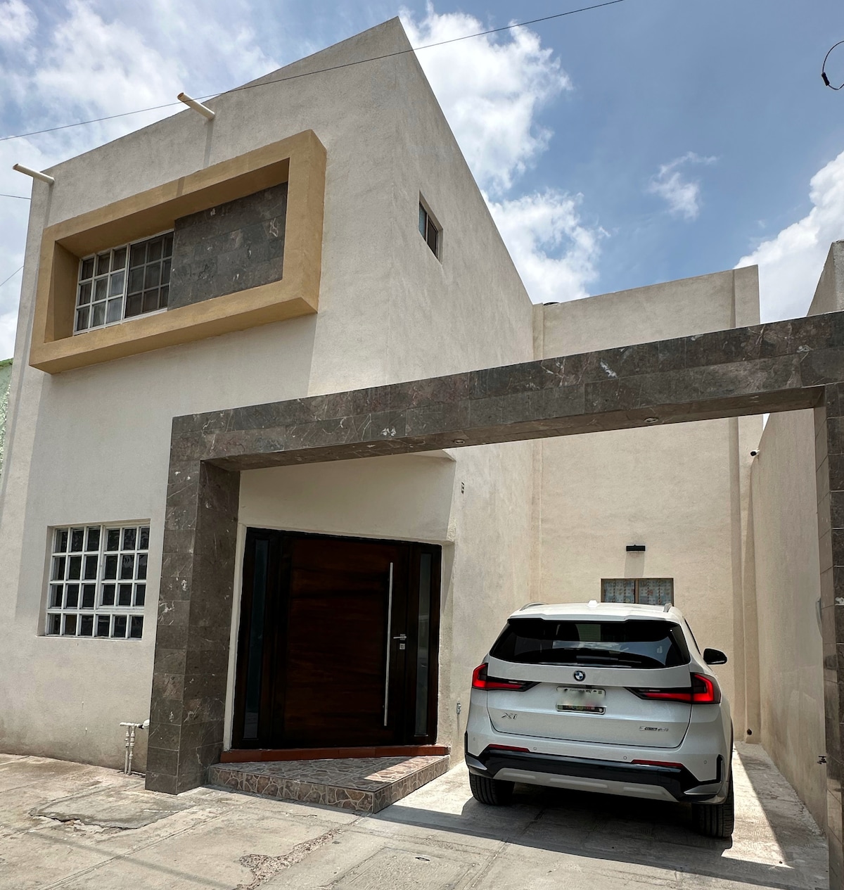 A modern residence is depicted with a two-story exterior featuring a combination of white and stone elements. A white vehicle is parked in the carport, and large windows provide natural light, enhancing the overall contemporary design.