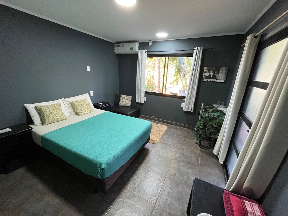 A spacious bedroom is presented with a large bed covered in a turquoise blanket. Natural light filters through two windows, illuminating the room's warm tones. A comfortable chair and a small side table are positioned beside the bed, enhancing the cozy environment.