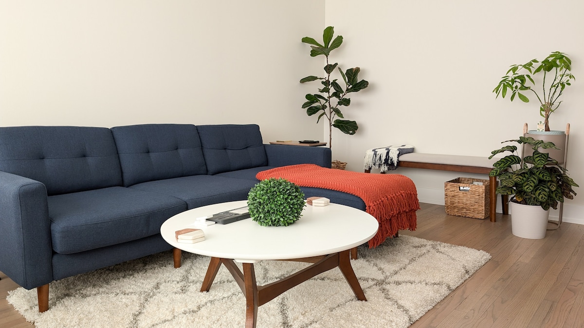 A spacious living area features a navy blue sectional sofa paired with a round white coffee table. Soft textures are added by a knitted red throw draped over the sofa. Potted plants stand by the walls, contributing to a refreshing ambiance.