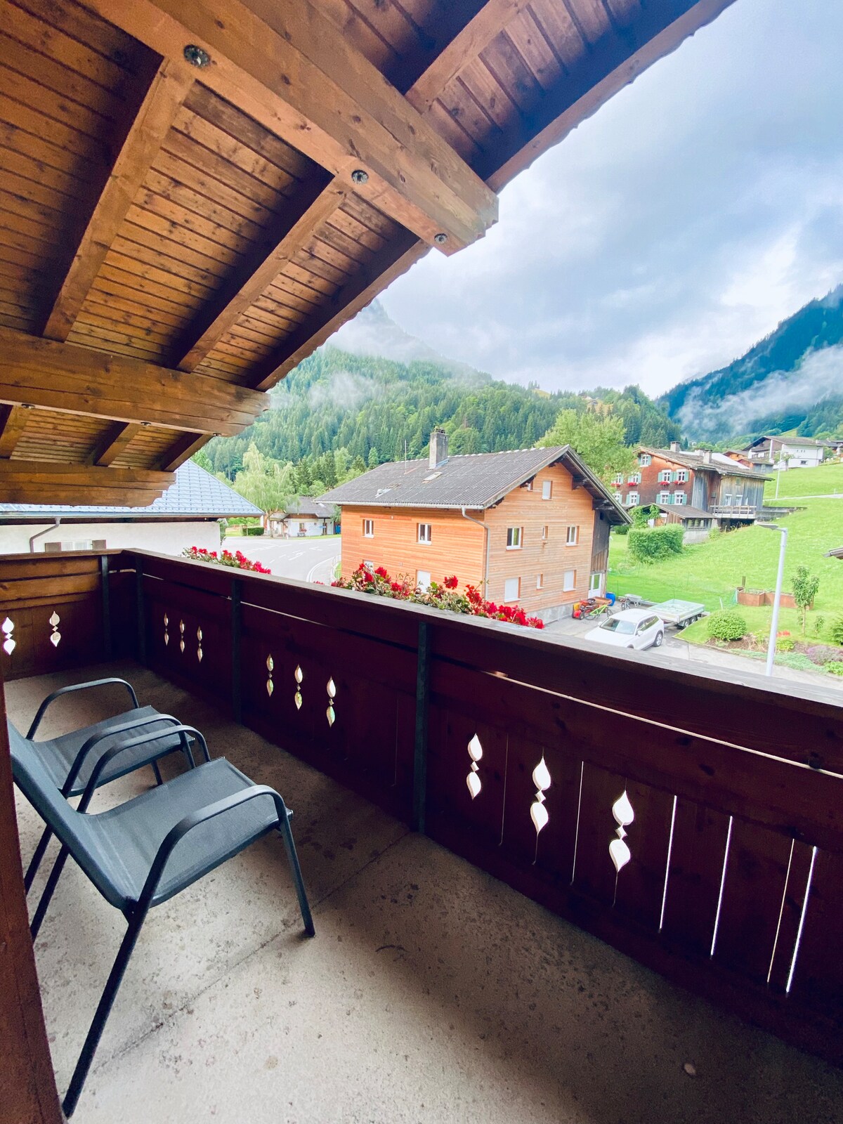 A private balcony features two simple chairs overlooking a picturesque view of the surrounding mountains and nearby houses. Red flowers line the balcony railing, enhancing the serene outdoor space that invites relaxation.