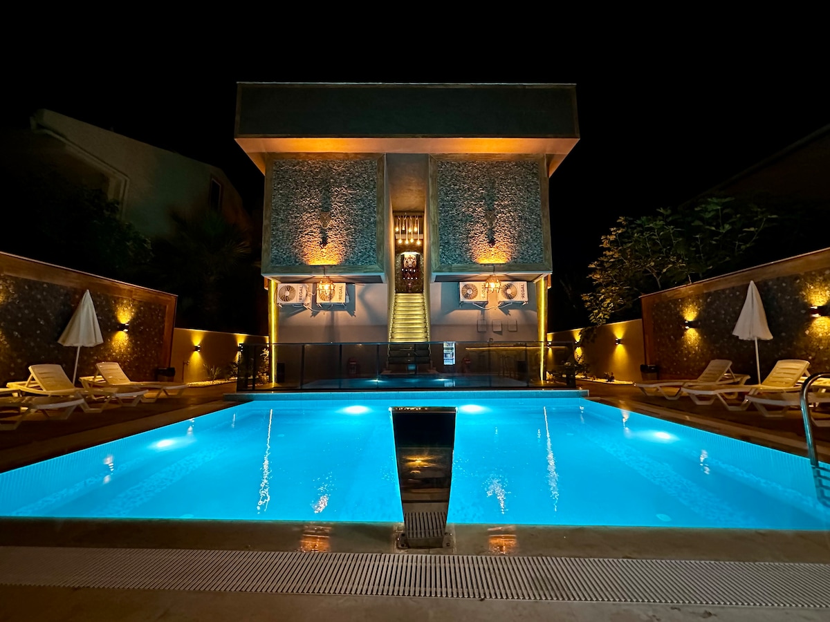 An outdoor swimming pool is illuminated at night with serene blue waters. The surrounding area features lounge chairs and white umbrellas, with a modern building in the background showcasing warm lighting, creating a calm atmosphere. Stairs lead up to the entrance, enhancing accessibility.