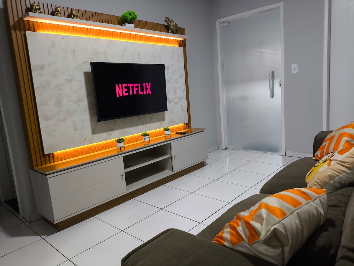 A modern living space features a spacious sofa with decorative pillows. A smart TV mounted on a stylish cabinet displays the Netflix logo. Soft ambient lighting highlights the cabinetry, and the tiled floor reflects the light, creating a welcoming atmosphere.