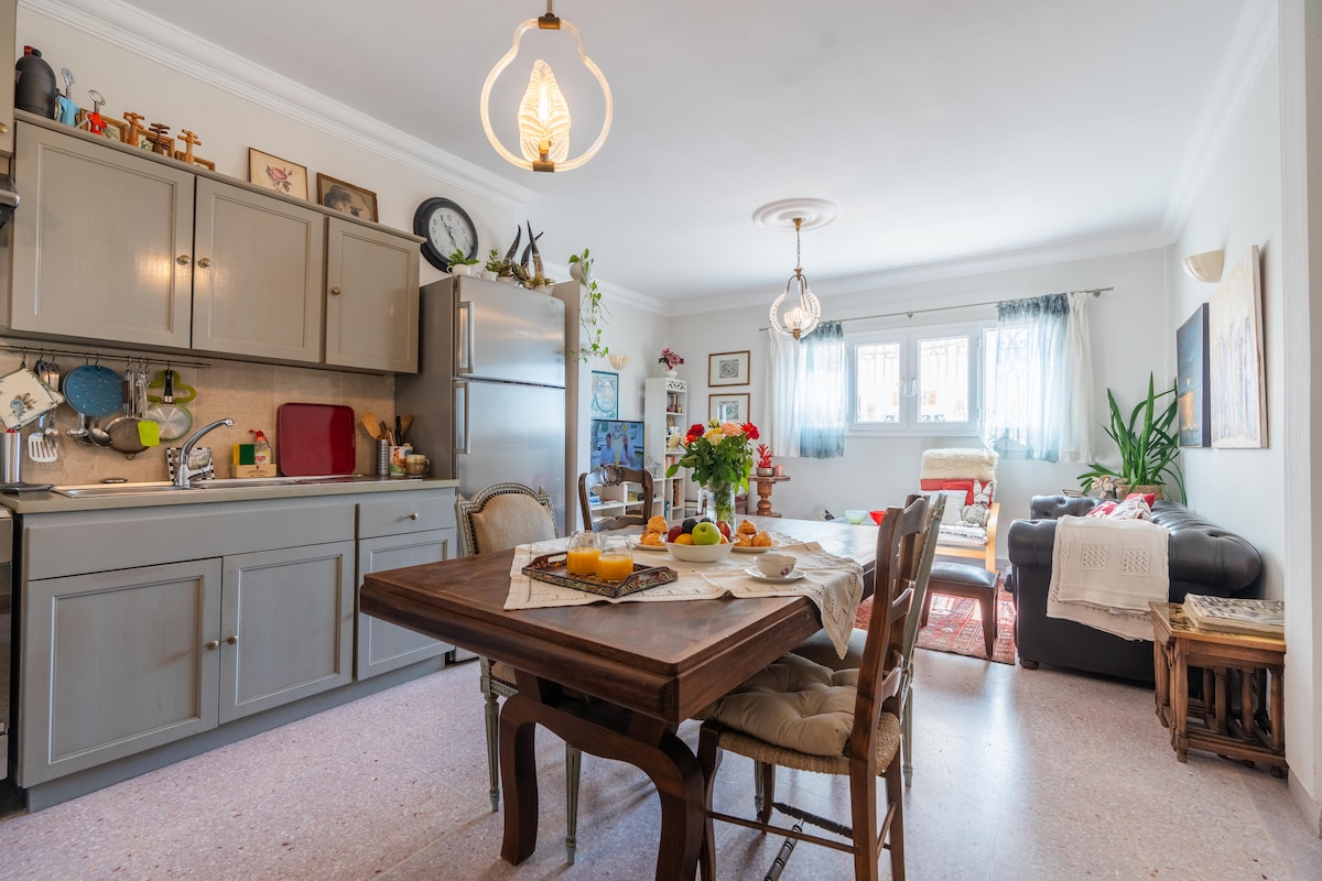 The kitchen and dining area feature a wooden table set for breakfast, accompanied by chairs. Cabinets line the walls, filled with kitchenware. A seating area is visible, with a couch and decorative plants enhancing the space. Light fixtures hang from the ceiling, providing illumination.