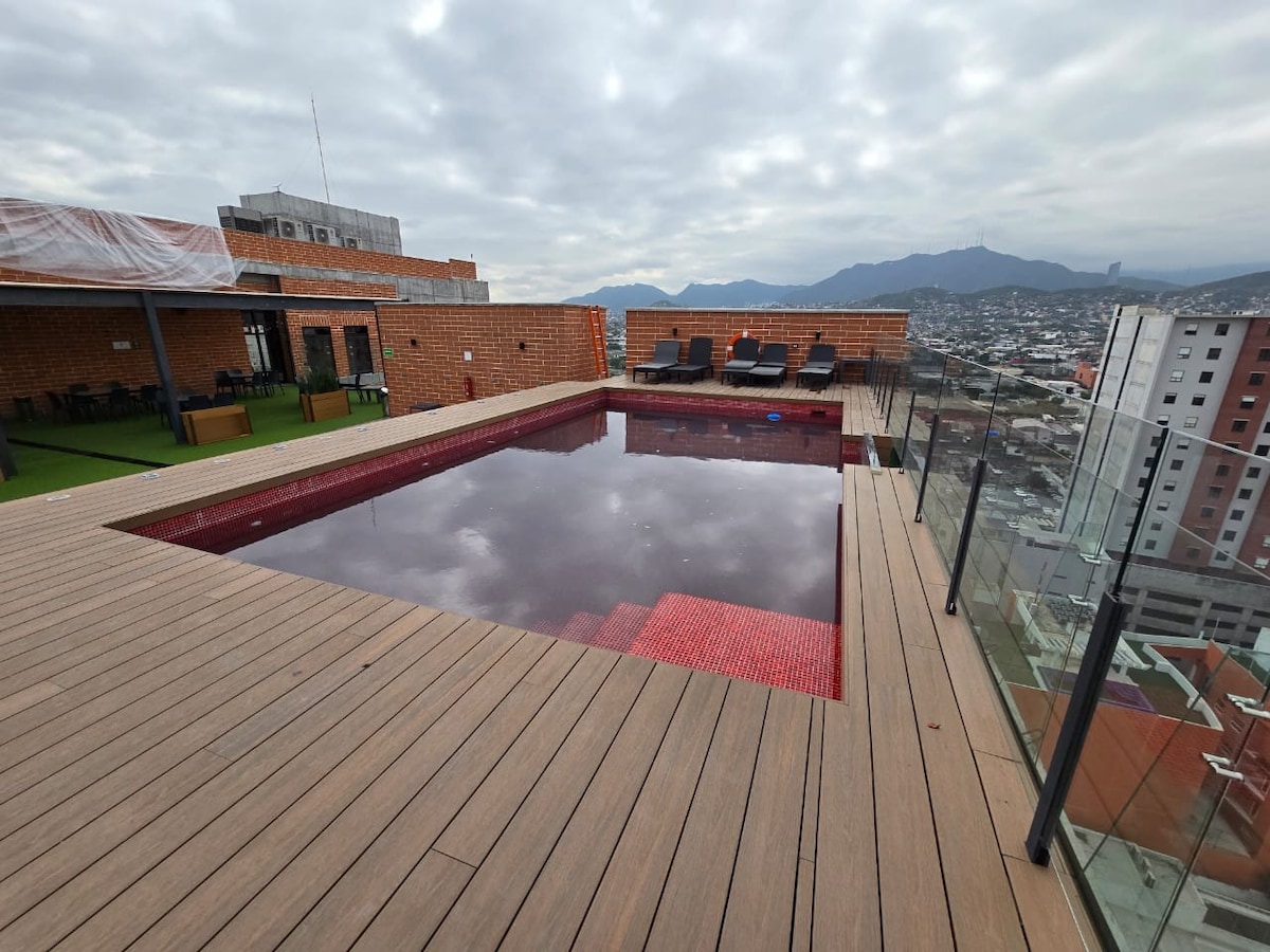 A rooftop pool is surrounded by a wooden deck, with multiple lounge chairs positioned nearby. The water reflects the cloudy sky and offers scenic views of the surrounding mountains and cityscape.