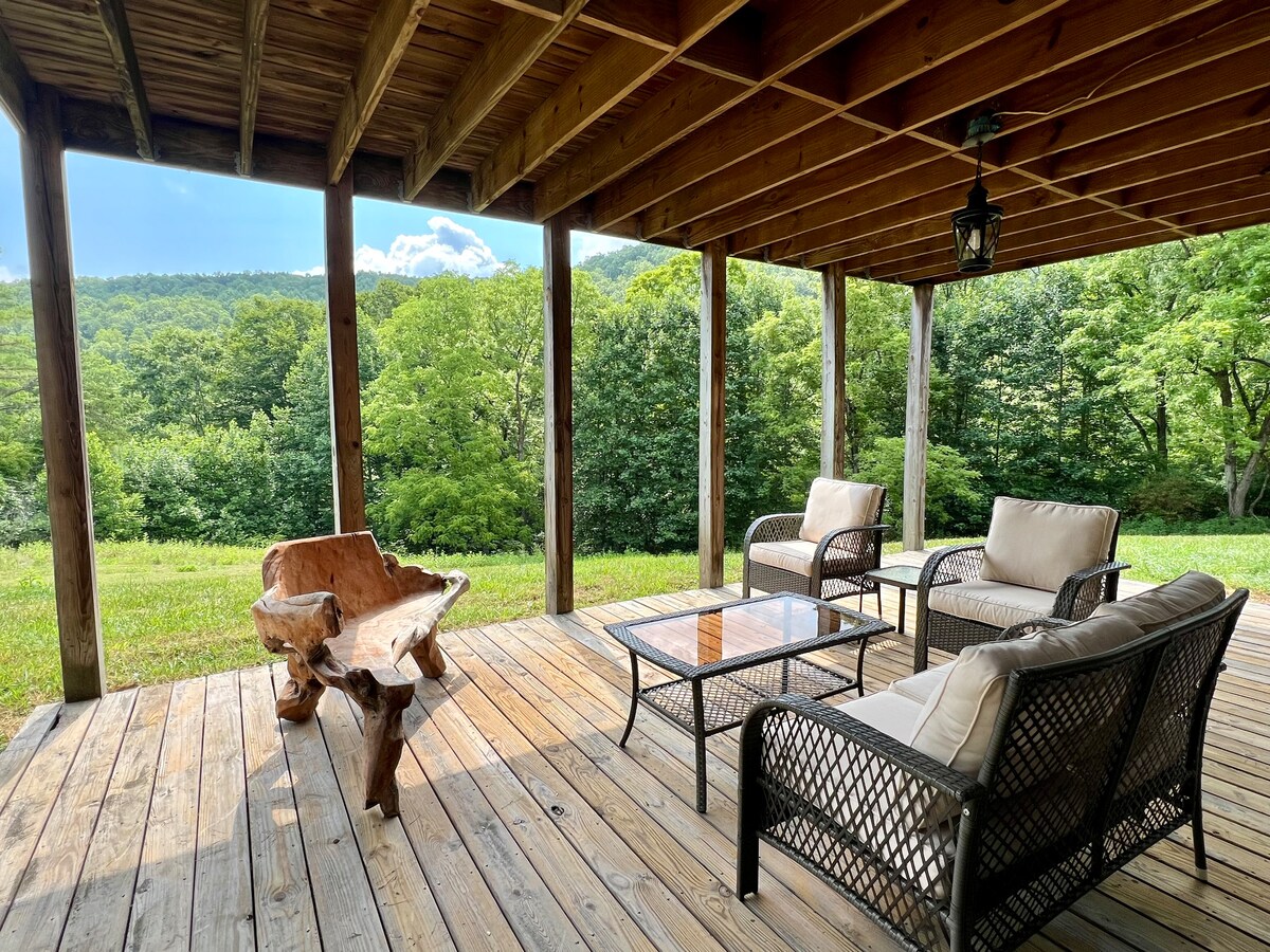 A covered porch extends the living space outdoors, featuring comfortable seating arrangements. A wooden coffee table is accompanied by a rustic accent chair and cushioned armchairs. Dense greenery and mountain views create a serene backdrop, enhancing the outdoor experience.