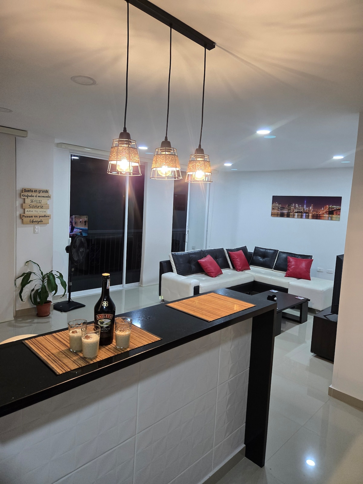 A modern living area is complemented by sleek furnishings, featuring a white sectional sofa with dark accents and vibrant red cushions. A stylish bar area with a black countertop is highlighted by pendant lighting, while large windows invite natural light and offer a view of the exterior.