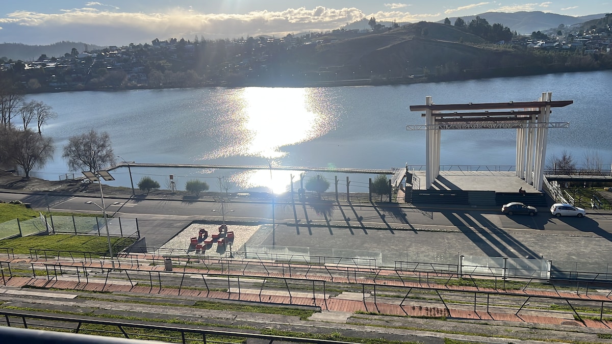 A scenic view of Laguna La Señoraza is presented, featuring a large, open area with a performance stage overlooking the water. Sunlight reflects on the lake's surface, while grassy spaces and pathways are visible in the foreground, encouraging leisurely strolls along the water's edge.