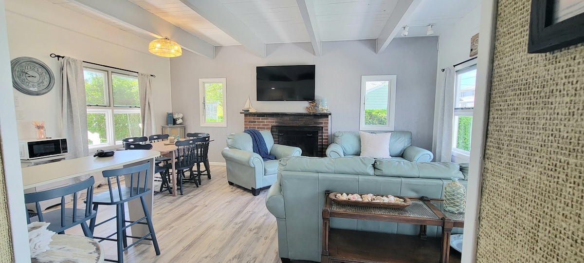 An open living area features a cozy seating arrangement with a light-colored couch and armchairs. Large windows allow natural light to fill the space, highlighting a decorative fireplace and a modern television mounted on the wall. A dining table is set nearby, conducive for gatherings.
