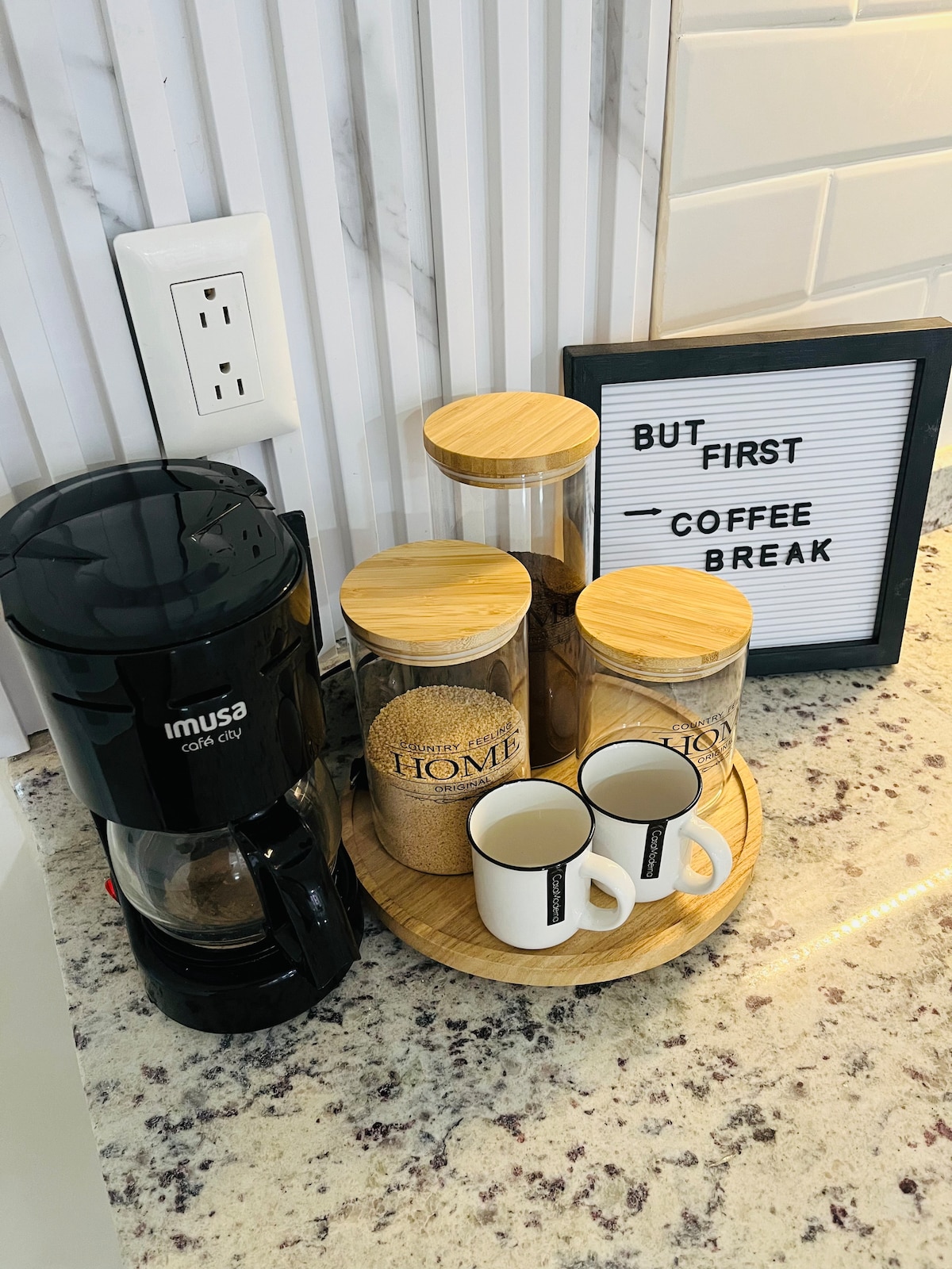 A coffee station is displayed, featuring a coffee maker, two small cups, and three storage jars with bamboo lids. The jars contain assorted ingredients for coffee. A sign reading 'But first - coffee break' adds a touch of warmth to the countertop with a marble surface.