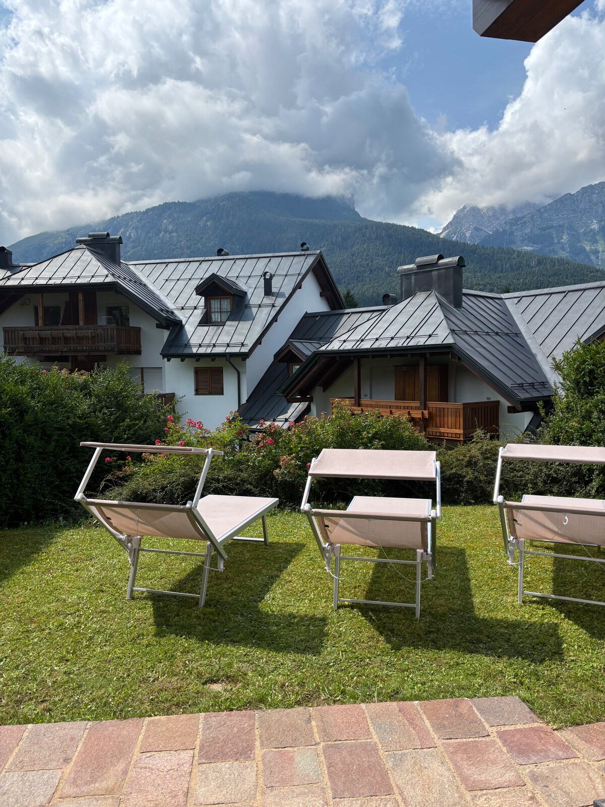A serene garden area is depicted, featuring several lounge chairs arranged neatly on a lush green lawn. The backdrop includes charming alpine-style buildings alongside majestic mountains, under a sky adorned with soft clouds.