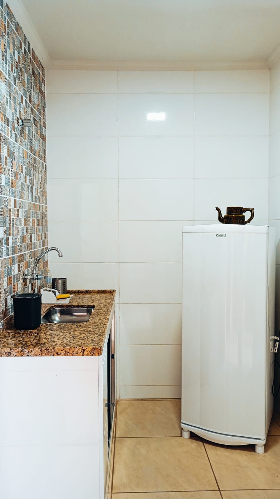 A compact kitchen features a combination of tiled and smooth white walls. A granite countertop hosts a small sink and cooking area. An upright refrigerator stands beside the countertop, while a decorative teapot adds a tasteful touch.