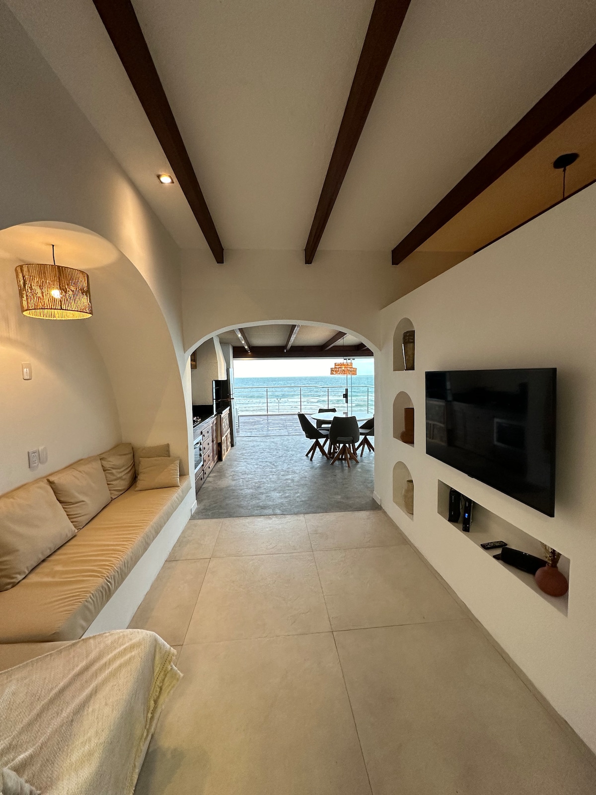 An open living area features a comfortable, neutral-toned sectional sofa positioned along a wall. A wall-mounted television is visible, and a dining area is seen through an archway, showcasing a table set for four with ocean views in the background.
