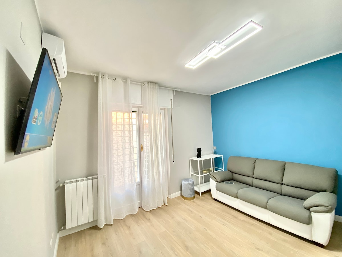 A bright living area showcases a gray sofa positioned against a blue accent wall. Light filters through sheer curtains, illuminating a flat-screen TV mounted on the wall. A modern white shelving unit and a small table complement the space, creating a comfortable environment.