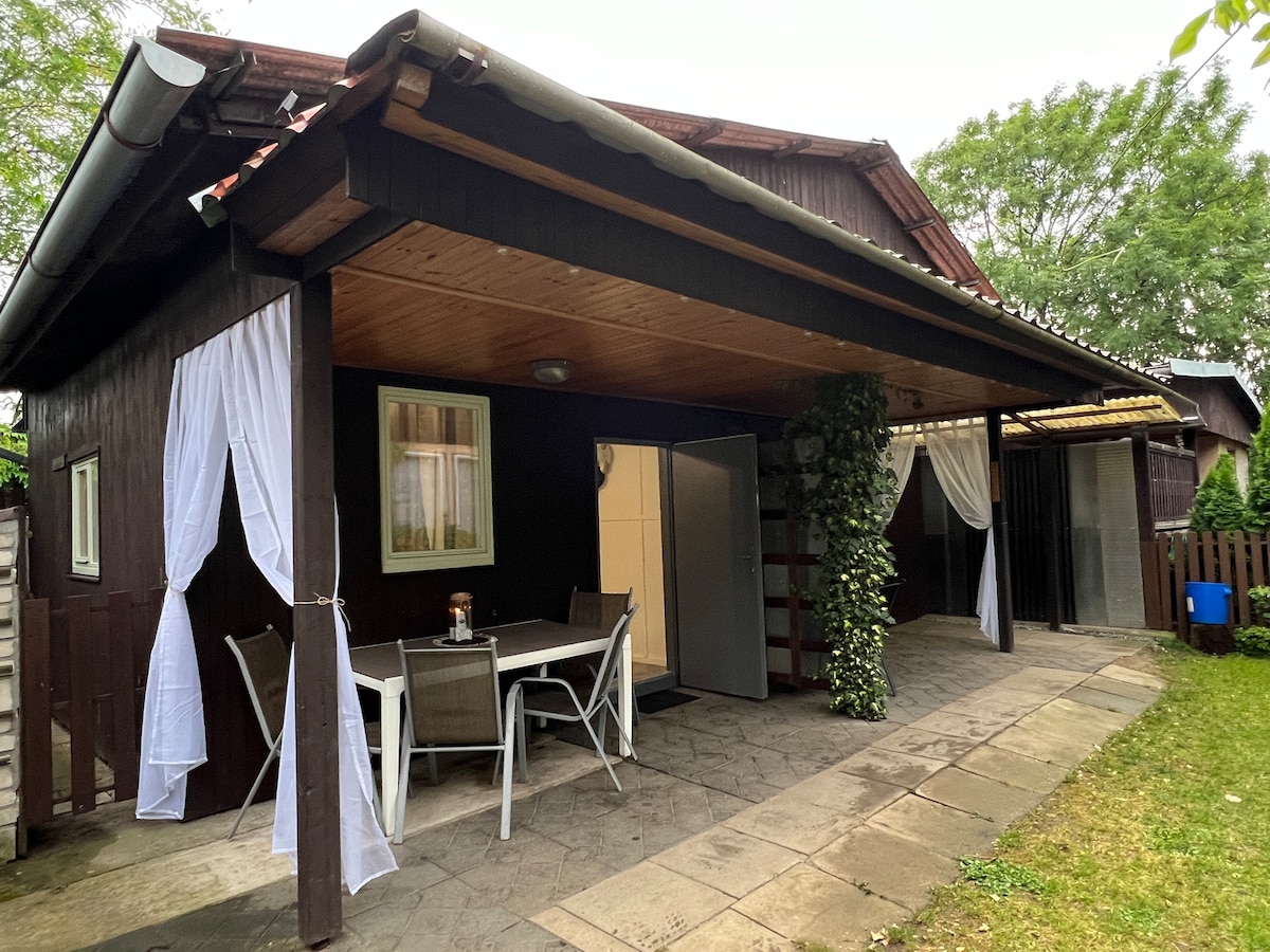 A shaded outdoor area features a table with four chairs, set against a backdrop of wooden walls. Draped white curtains add a soft touch, while the grassy space surrounding the patio provides a natural setting. An entryway leads to indoor areas.