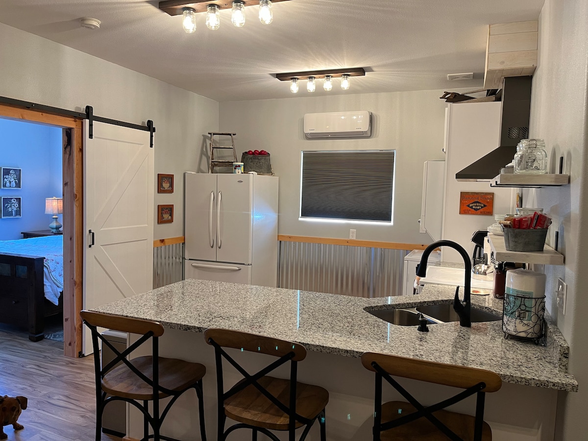 An open kitchen showcases modern appliances against a backdrop of light cabinetry and granite countertops. Three wooden stools are situated at the breakfast bar, while an air conditioning unit is mounted above. A sliding barn door leads to another room, enhancing the space's character.
