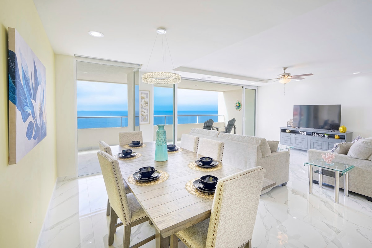 A bright and airy dining area features a large wooden table set for six, complemented by elegant dishware. Large glass doors showcase expansive ocean views, while the open layout connects to a comfortable living space, highlighted by a light-colored sofa and a wall-mounted television.