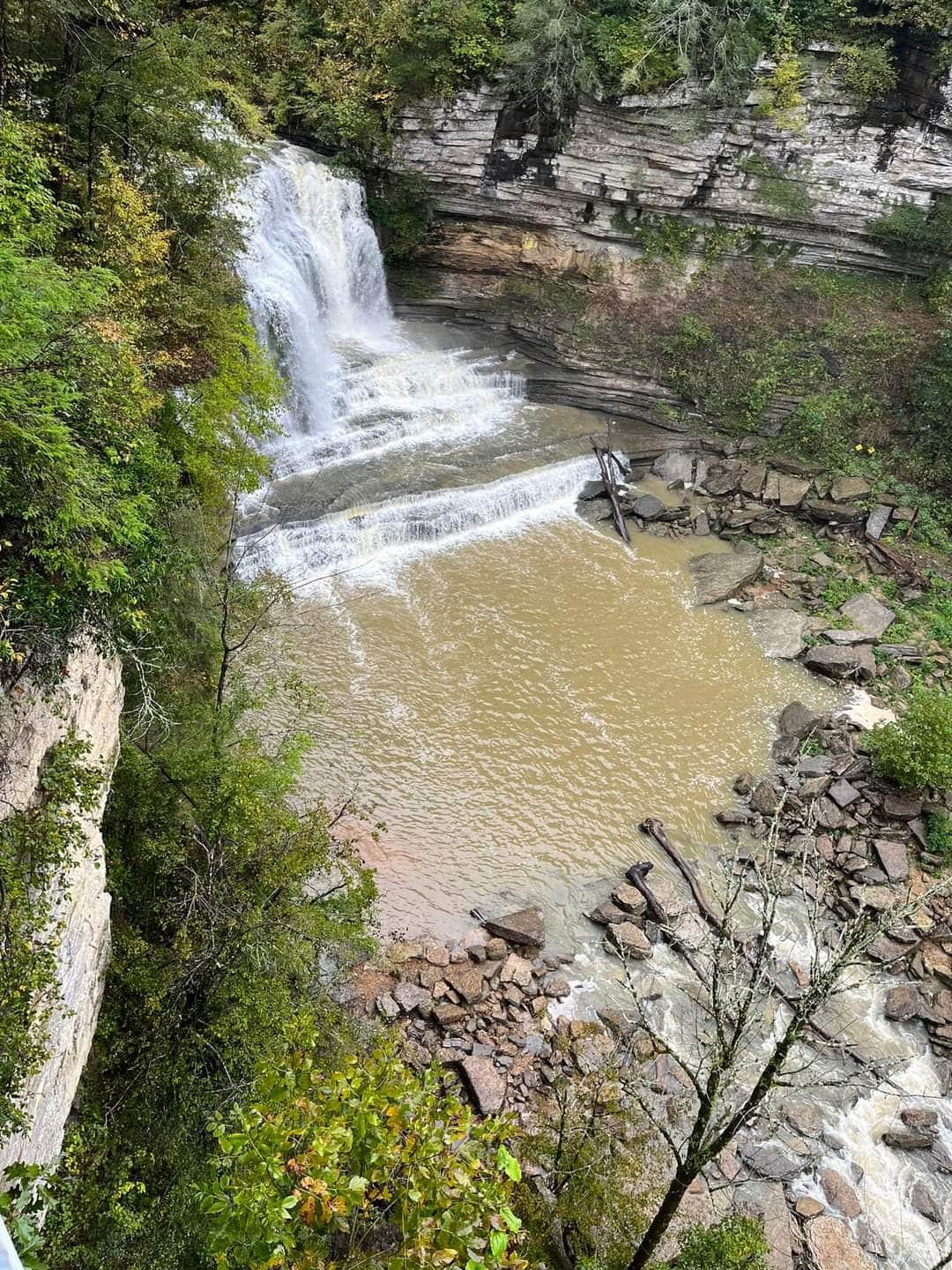 A cascading waterfall flows into a serene pool, surrounded by rugged stone walls and foliage. The water tumbles over rocky ledges, creating a natural backdrop of earthy tones and greenery. The landscape reflects the tranquility of the rural setting.