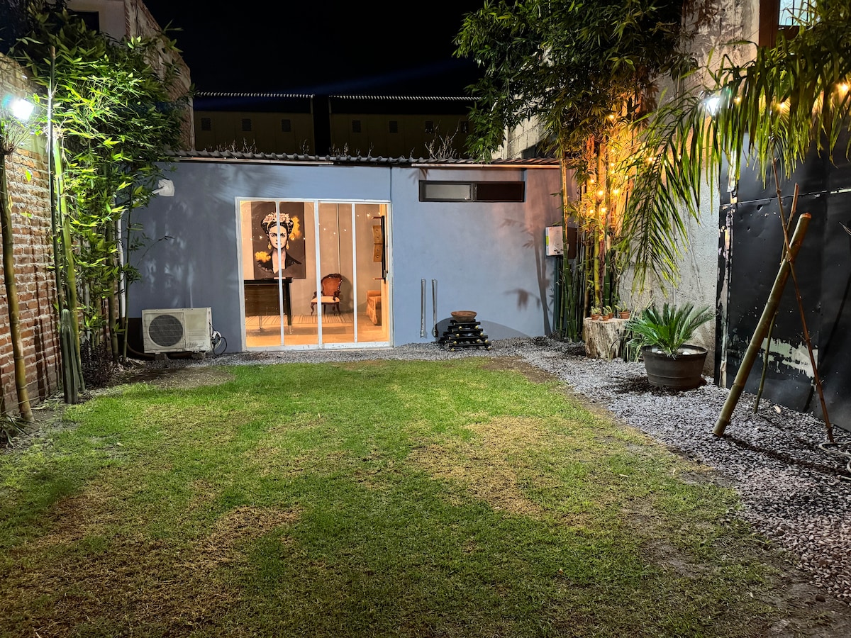A tranquil garden area is featured, illuminated softly by outdoor lighting. Green grass is bordered by pebbles and plants, while a wall is adorned with climbing foliage. Glass doors at the far end reveal an interior space, enhancing the connection between indoor and outdoor environments.