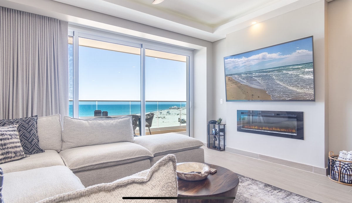 A spacious living area features a large sectional sofa, with a sleek coffee table in front. A mounted television displays a serene beach scene. Floor-to-ceiling windows provide unobstructed views of the ocean and beach, allowing natural light to fill the room.