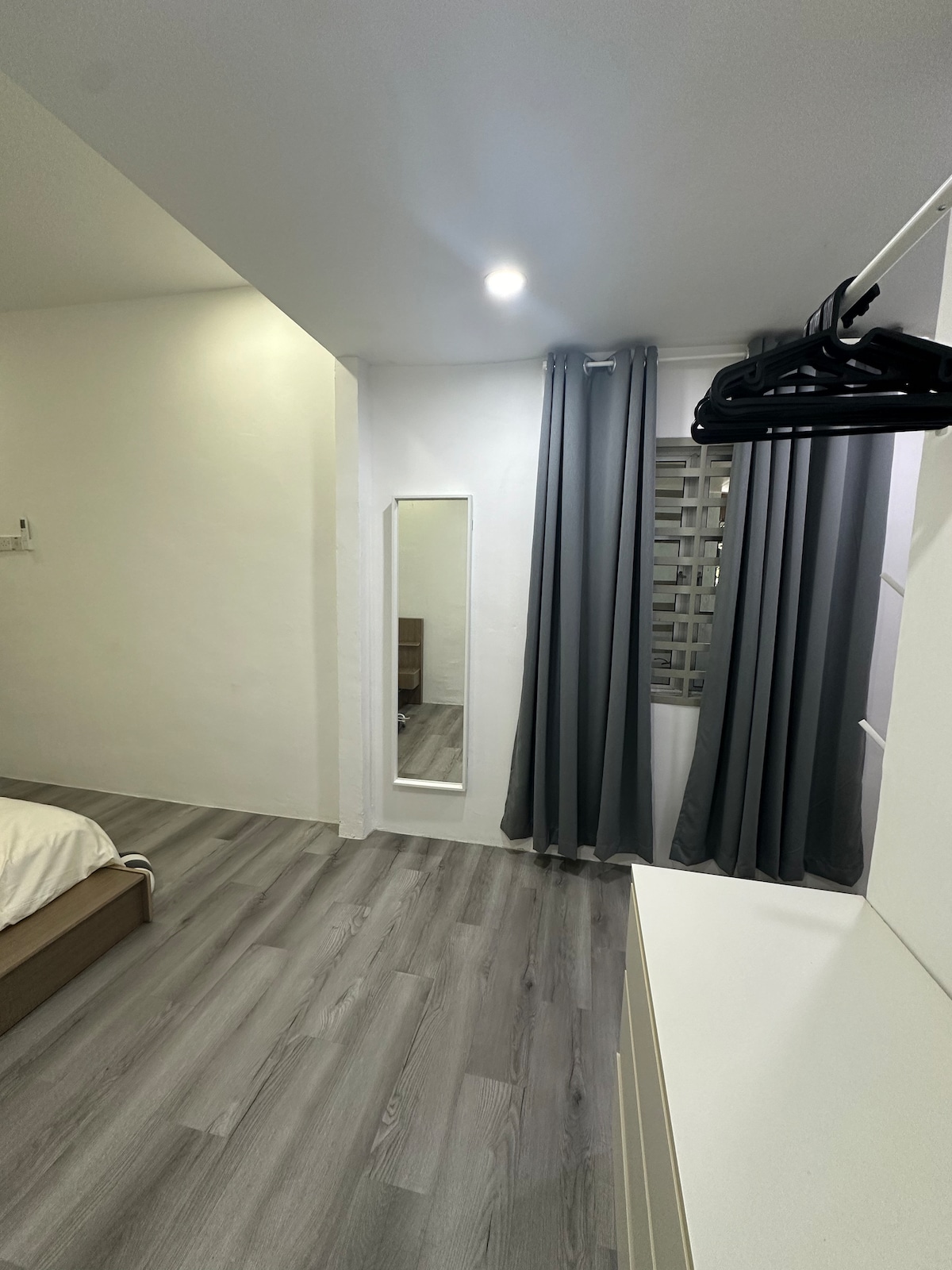 A bright room features light grey walls and a sleek floor. A full-length mirror is positioned against the wall, complemented by curtains that frame a window. A spacious bed is dressed in neutral linens, and a few hangers are visible on a minimalist rack.