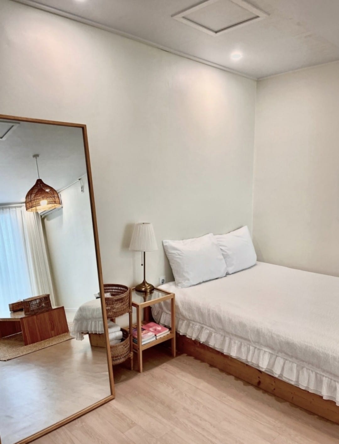 A well-lit sleeping area is presented with a single bed dressed in white linens. A tall mirror leans against the wall, reflecting the space. A bedside table holds a lamp and a small stack of magazines. Natural light filters in through nearby curtains.