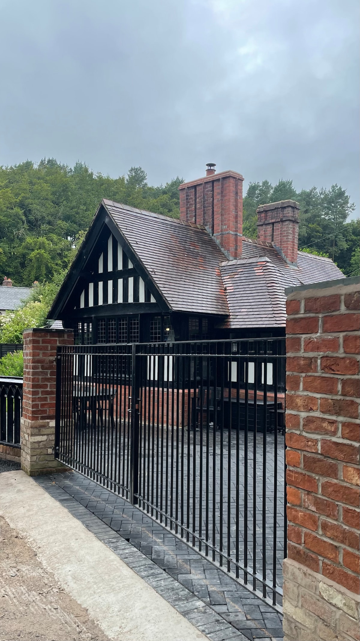 A historic lodge with half-timbered architecture features a steeply pitched roof and prominent chimneys. The entrance is framed by a black metal gate, and a paved driveway leads to the front porch. Lush greenery surrounds the property, contributing to a serene atmosphere.