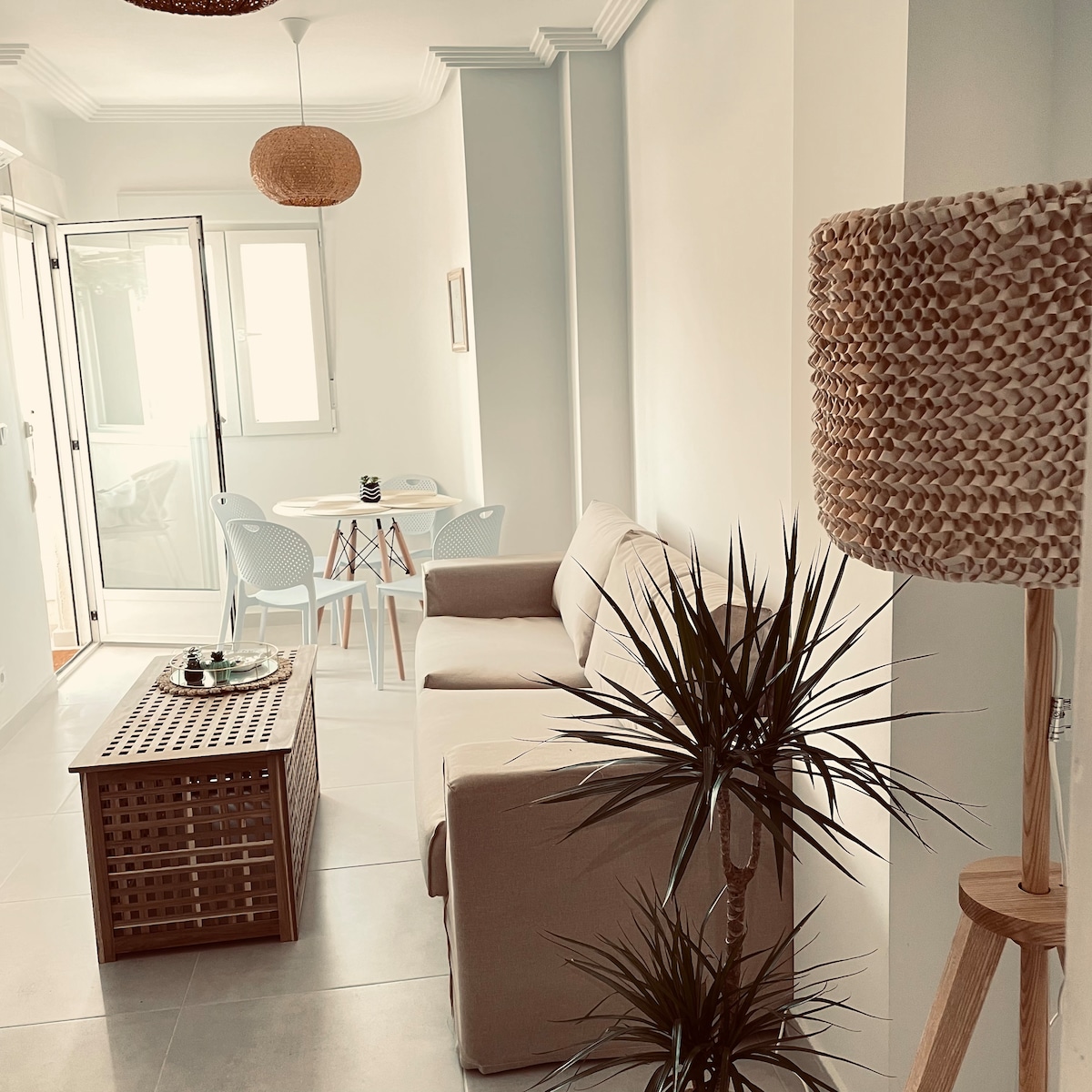 A light-filled living area is presented, featuring a beige sofa and a wooden coffee table. A dining set with two chairs stands beside a window, while a tall plant adds greenery to the space. A woven lamp provides soft lighting, enhancing the room's ambiance.